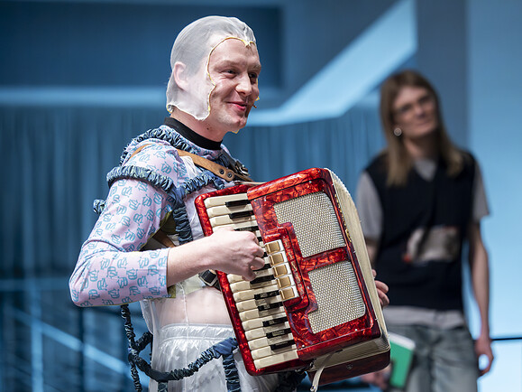 Ghost spielt rotes Akkordeon, trägt Haube und Kostüm, lächelt, im Hintergrund steht eine weitere Person.-177163