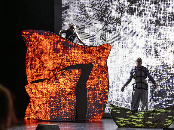 Teen steht oben links auf Kissenkulisse, Ghost unten rechts. Kulisse rot beleuchtet, Hintergrund hell, Bühne bedrohlich.-17ea76