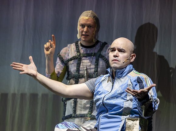 Parent sitzt mit offenen Armen, genervter Blick. Ghost dahinter zeigt nach oben. Beide tragen Kostüme, Bühnenvorhang im Hintergrund.-30c7e7