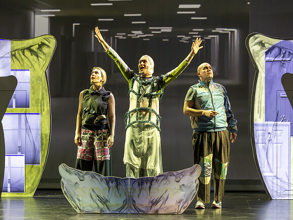 Teen, Ghost, Parent stehen im Bühnenbild. Im Hintergrund Projektion von einem Raum, vorne große Kissenkulisse, Ghost hebt Arme.-49f6b1