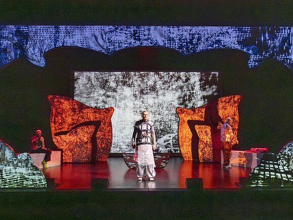 Große Bühne mit Teen, Ghost, Parent und großem Bildschirm im Hintergrund. Bühnenbild leuchtet rot und schwarz, Personen stehen und sitzen verteilt auf der Bühne.-80508b