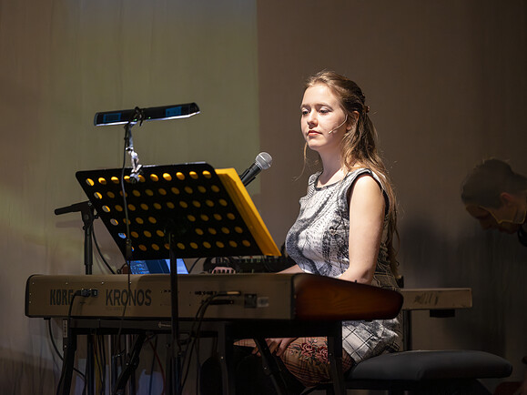 ViKI sitzt mit Mikrofon am Keyboard auf einer Bühne. Sie trägt ein Kleid, vor ihr Notenständer, warmes Licht, im Hintergrund Bühne und Technik.-924e9d
