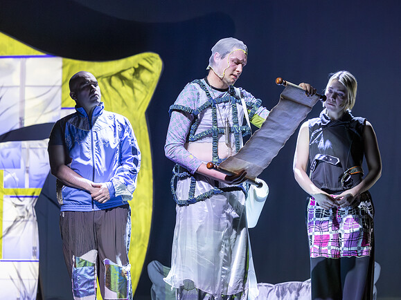 Parent, Ghost, Teen stehen auf Bühne, Ghost liest Schriftrolle. Links Fensterprojektion.-a9e92d