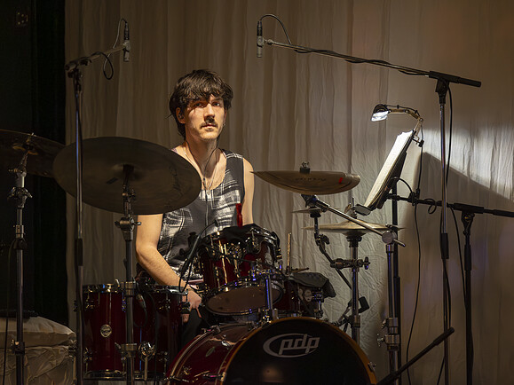 Mann sitzt am Schlagzeug auf einer Bühne. Braune Haare, ärmelloses Shirt, Mikrofone über dem Schlagzeug, Vorhang im Hintergrund.-bcc53d