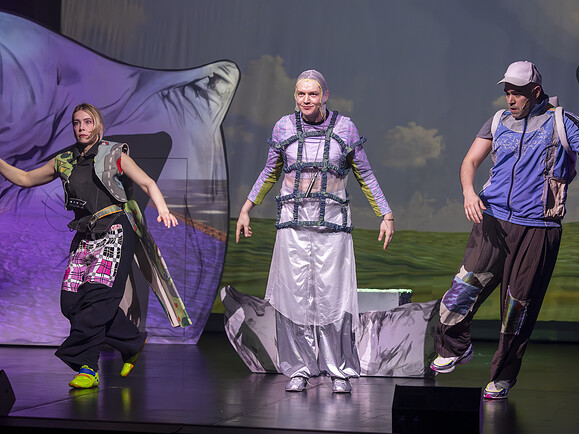 Teen, Ghost, Parent stehen nebeneinander auf Bühne. Sie tragen bunte Kostüme, hinter ihnen großes Bühnenbild mit Landschaft.-cb2690