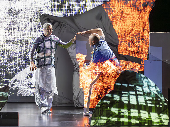 Ghost und Parent im Bühnenbild mit Projektion. Parent klammert an Kissenkulisse, Ghost schaut von links, rotes und weißes Licht.-e90f3f